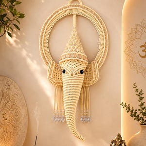 May include: A cream-colored macrame wall hanging shaped like the head of Ganesha, featuring a circular frame, intricate knotted patterns, a pointed crown, large ears, and a trunk. The design includes dangling clear crystal beads at the base.