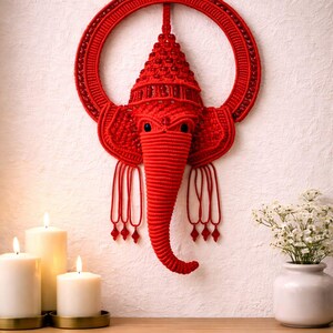 May include: A red macrame wall hanging shaped like the head of an elephant with a trunk, large ears, and a circular frame. The design features intricate knotted cord work, embedded red beads, and dangling cord tassels with small red bead accents.