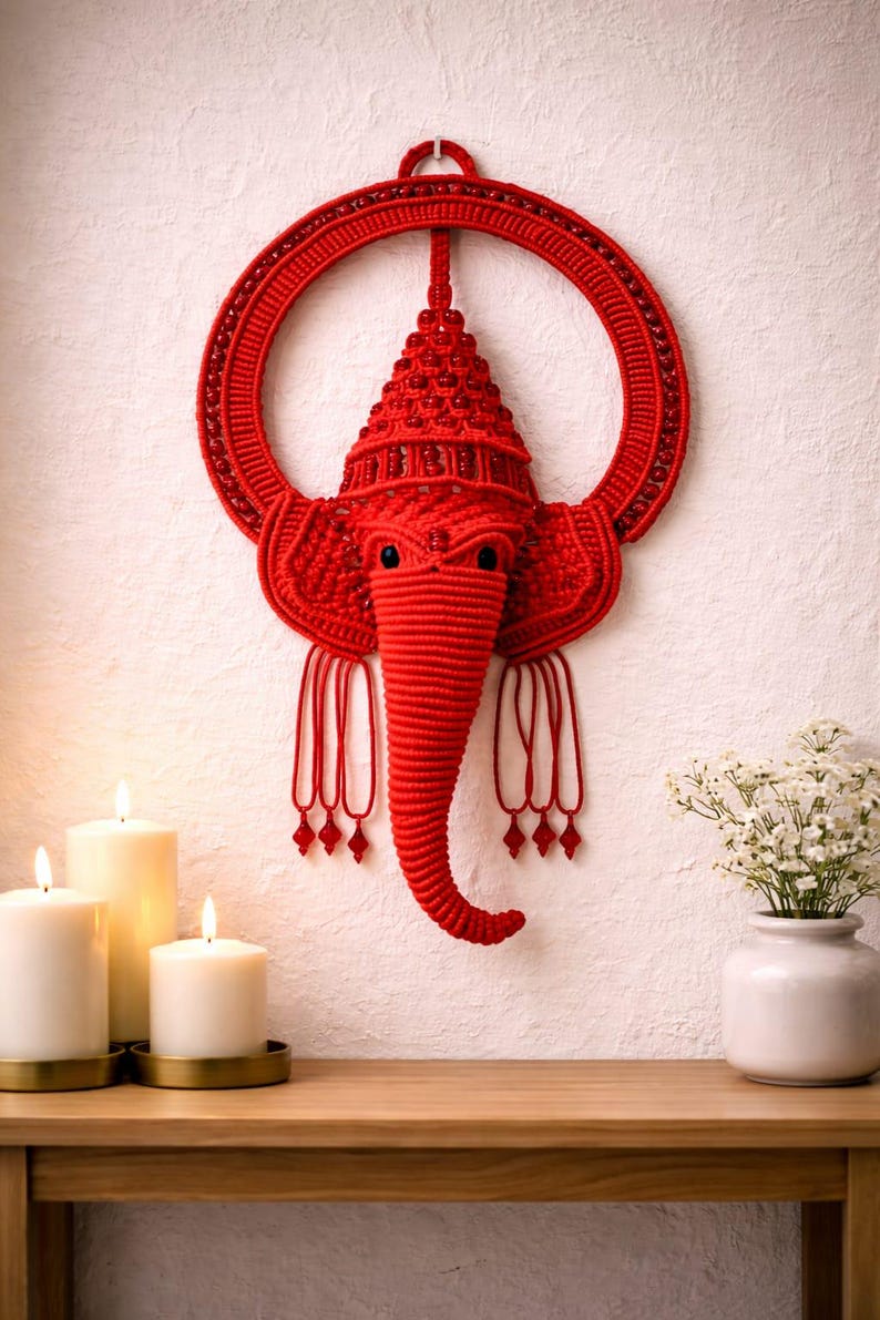 May include: A red macrame wall hanging shaped like the head of an elephant with a trunk, large ears, and a circular frame. The design features intricate knotted cord work, embedded red beads, and dangling cord tassels with small red bead accents.
