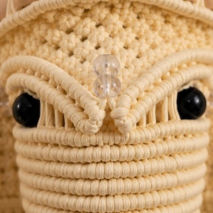 May include: A macrame wall hanging crafted from cream-colored cord, featuring a detailed woven pattern with smooth beige wooden beads along the top edge, two clear glass beads in the center, and two large, round, glossy black beads for eyes.