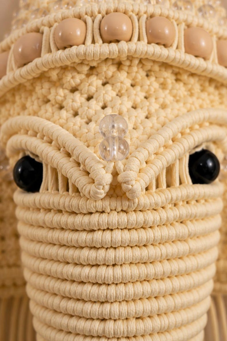 May include: A macrame wall hanging crafted from cream-colored cord, featuring a detailed woven pattern with smooth beige wooden beads along the top edge, two clear glass beads in the center, and two large, round, glossy black beads for eyes.