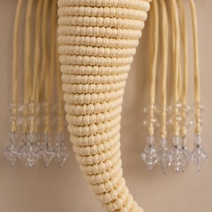 May include: A cream-colored macrame wall hanging shaped like an elephant head. The trunk is made of tightly knotted cord, and the sides feature long, dangling cords finished with clear, faceted, diamond-shaped acrylic beads at the ends.