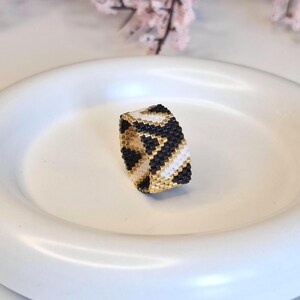 May include: A beaded ring with a geometric pattern in black, white, and gold. The ring is set on a white plate, showcasing the intricate beadwork and color scheme. The ring's design features a repeating chevron pattern.