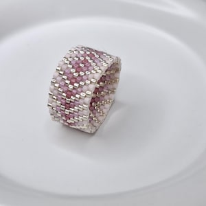 May include: A wide band ring crafted from small, cylindrical beads in shades of light pink, dark pink, and metallic silver. The beads are arranged in a repeating chevron pattern that wraps around the entire circumference of the ring.