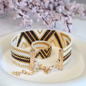 May include: A beaded bracelet and ring set with a chevron pattern in white, brown, and gold. The bracelet has a gold clasp and chain. The jewelry is displayed on a white plate with a blurred floral background.