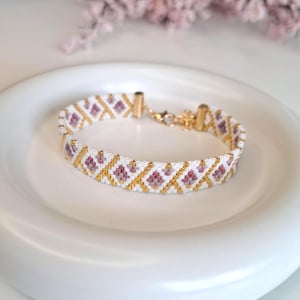 Puede incluir: Una pulsera de cuentas sobre un fondo blanco, con un patrón repetido de cuentas doradas, blancas y moradas. La pulsera se asegura con un cierre dorado. La pulsera se muestra en un plato blanco.