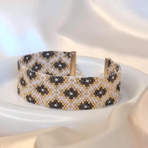 Op de afbeelding: Een geweven kralenarmband met een herhalend ruitpatroon in wit, metallic goud en donker antracietgrijs. De armband is afgewerkt met twee cilindrische goudkleurige metalen eindkappen en een delicaat kettingmechanisme als sluiting aan de achterkant.