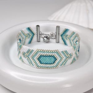May include: A woven bead bracelet featuring a geometric pattern in white, light blue, and metallic silver. The bracelet is constructed from small, uniform beads and includes a silver-toned metal clasp mechanism with a small dangling charm.