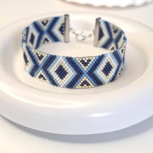 May include: A woven bead bracelet featuring a geometric pattern of diamonds and triangles in shades of navy blue, light blue, white, and metallic gold. The bracelet is finished with silver-toned metal end caps and a lobster claw clasp closure.