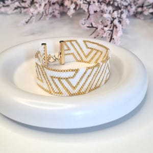 May include: A white and gold beaded bracelet with a chevron pattern. The bracelet is made of small, white beads with gold accents, and features a gold clasp. The bracelet is displayed on a white, round dish.