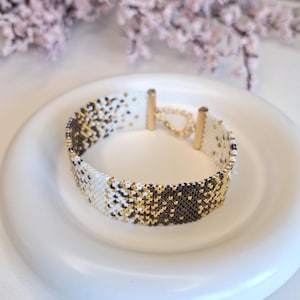Può includere: Un braccialetto di perline con un motivo geometrico in bianco, nero e oro. Il braccialetto è su un piatto bianco e ha chiusure color oro.