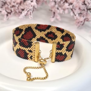 May include: A beaded bracelet with a leopard print design. The bracelet features a gold, black, and burgundy pattern. The adjustable closure is gold-toned. The bracelet is on a white plate.
