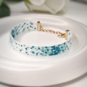 May include: A woven beaded bracelet featuring a pixelated pattern of white, light blue, and dark teal beads. The bracelet is finished with gold-toned metal end caps and a gold-toned metal chain extender with a lobster claw clasp closure.