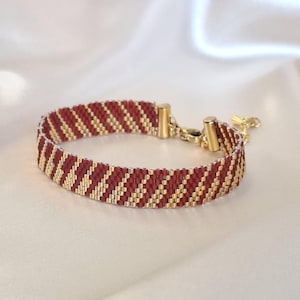 May include: A woven beaded bracelet featuring a repeating diagonal pattern of matte maroon and metallic gold seed beads. The bracelet is finished with gold-toned metal end caps, a lobster claw clasp, and a short extender chain with a small charm.