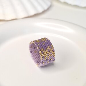 Peut inclure: Une bague large composée de petites perles carrées disposées selon un motif répétitif alternant le lilas doux et l'or métallique. Le perlage forme une grille dense et uniforme, créant une surface texturée sur toute la circonférence.