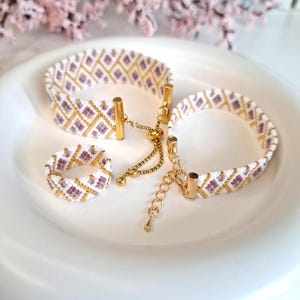May include: A jewellery set comprising two bracelets and a ring, all beaded. The pieces display a white base with a repeating pattern of gold and purple diamond shapes. The bracelets have gold-coloured clasps and chains.