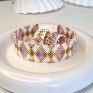 May include: A beaded cuff bracelet and a matching ring featuring a repeating diamond pattern in white, soft pink, and olive green, accented with metallic gold beads. The jewelry set is constructed from small, uniform glass beads in a woven design.
