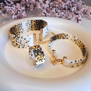 Könnte beinhalten: Ein Perlen-Schmuckset mit einem Armband, einem Ring und einem weiteren Armband. Die Teile sind weiß mit schwarzen und goldenen Akzenten. Das Armband hat eine goldene Kette und einen Verschluss. Der Ring ist quadratisch. Der Schmuck wird auf einem weißen Teller präsentiert.