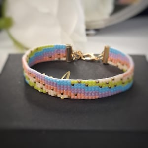 Könnte beinhalten: Ein Perlenarmband mit einem sich wiederholenden Wellenmuster in Pastelltönen wie Hellblau, Lavendel, Pfirsich und Limettengrün. Das Armband ist mit goldfarbenen Metallendkappen und einem passenden Karabinerverschluss versehen.