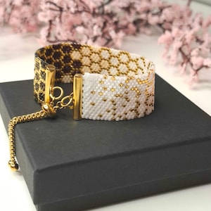 May include: A beaded bracelet with a gold chain and clasp. The bracelet features a geometric pattern in black, white, and gold. The bracelet is resting on a black box, with pink flowers in the background.
