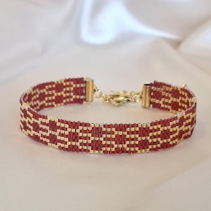 Op de afbeelding: Een geweven kralenarmband met een herhalend geometrisch patroon in dieprood en metallic goud. De armband is afgewerkt met goudkleurige metalen eindkappen en een karabijnsluiting, wat een gestructureerde band van kleine glaskralen vormt.