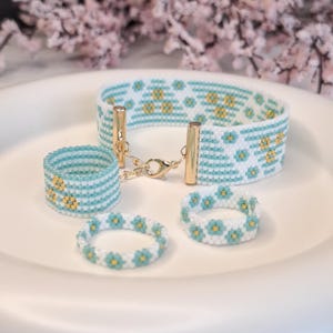 May include: A beaded jewelry set featuring a bracelet and three rings. The bracelet has gold-colored metal clasps and a pattern of white, light blue, and gold beads. The rings have similar patterns, with a floral design. The jewelry is displayed on a white surface.