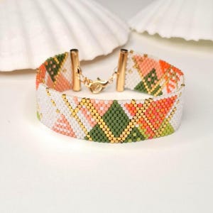 May include: A woven bead bracelet featuring a geometric pattern of white, olive green, peach, and bright orange beads. The bracelet is accented with small gold-colored beads and finished with gold-toned metal end caps and a lobster claw clasp.
