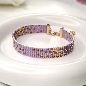 Puede incluir: Una pulsera de cuentas hecha de pequeñas cuentas cuadradas en tonos morado claro, morado oscuro y oro metálico. La pulsera presenta un diseño tejido plano con terminales de metal dorado y un mecanismo de cierre de tipo mosquetón.