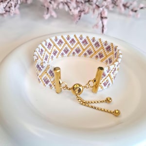 May include: A beaded bracelet with a white background, featuring a geometric pattern of purple and gold beads. The bracelet has gold-colored metal accents and a sliding clasp for adjustable sizing.
