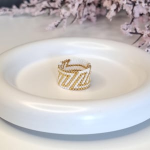 Könnte beinhalten: Ein Perlenring mit einem weißen und goldenen Zickzackmuster. Der Ring besteht aus kleinen, runden Perlen und hat eine Bandbreite von etwa 2 cm. Der Ring wird auf einer weißen, flachen Schale präsentiert.