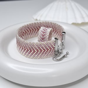 Puede incluir: Un conjunto de pulsera y anillo de cuentas con un patrón de chevrón en tonos rosa, malva y plata metálica. La pulsera incluye un mecanismo de cierre deslizante de metal plateado con una cadena delicada y pequeñas cuentas esféricas.