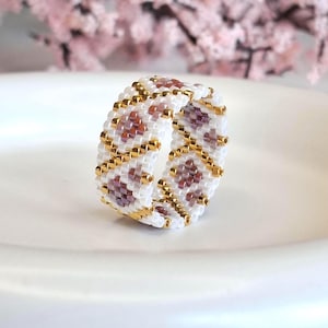 Puede incluir: Un anillo de cuentas con un diseño geométrico de cuentas blancas, doradas y burdeos. El anillo está hecho de pequeñas cuentas espaciadas uniformemente, creando una superficie texturizada. El anillo está sobre un fondo blanco, con flores rosas suaves en el fondo.