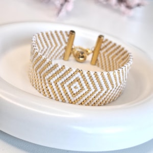 Könnte beinhalten: Armband aus weißen und goldenen Perlen mit geometrischem Muster. Das Armband hat einen goldenen Verschluss und wird auf einem weißen Teller präsentiert. Das Design beinhaltet eine zentrale Raute. Die Breite des Armbands beträgt ca. 2 cm.