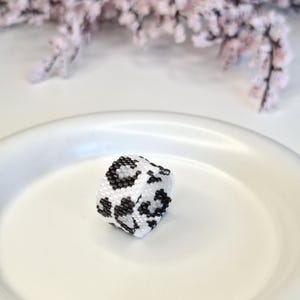 May include: A beaded ring with a white base and a black leopard print pattern. The ring is set against a white background, with a blurred floral arrangement in the background.