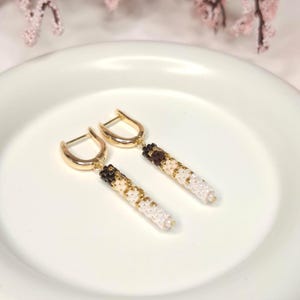 May include: A pair of gold-tone hoop earrings featuring dangling cylindrical pendants crafted from small beads in a gradient pattern. The beads transition from dark brown at the top to light beige and white at the bottom, creating a subtle ombre effect.