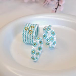 May include: Three beaded rings in different sizes. The rings have a pattern of white, light blue, and gold beads. The rings are displayed on a white, slightly curved surface. The rings are handmade and suitable for everyday wear.