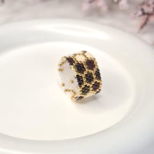 May include: A handmade beaded ring with a checkered pattern. The ring features a combination of black, gold, and cream-colored beads. The ring is set against a white background, highlighting the intricate beadwork and design.