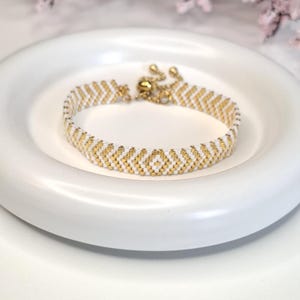 May include: A beaded bracelet with a white and gold chevron pattern. The bracelet is made of small, round beads and has a gold-colored adjustable clasp. The bracelet is displayed on a white, round plate.