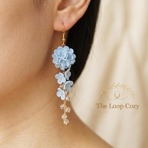 May include: A close-up of a light blue floral earring with a gold hook. The earring features a cluster of crocheted flowers and dangling clear beads. The Loop Cozy logo is visible in the background.
