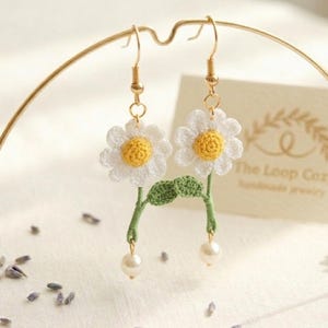 Puede incluir: Pendientes de crochet con forma de margarita, con pétalos blancos y centros amarillos. Los pendientes presentan tallos y hojas verdes, con una perla en la parte inferior de cada tallo. Se muestran en un expositor dorado.