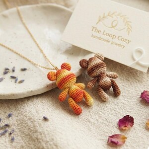 May include: Two crocheted teddy bear pendants, one orange and yellow striped, the other brown and tan striped, are displayed. A gold chain necklace is attached to the orange bear. A card with the text "The Loop Cozy handmade jewelry" is also visible.