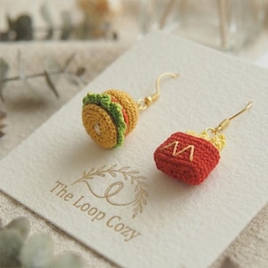 Può includere: Orecchini a tema cibo all'uncinetto. Uno è un hamburger con dettagli gialli, verdi e arancioni. L'altro è un contenitore rosso di patatine fritte con patatine gialle e un design dorato con una "M". Il logo The Loop Cozy è visibile.