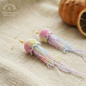 Può includere: Un paio di orecchini a forma di medusa fatti a mano. Gli orecchini presentano un design a medusa all'uncinetto con un arcobaleno di colori, tra cui rosa, giallo e blu. Gli orecchini hanno ganci color oro e il testo "The Loop Cozy handmade jewelry".