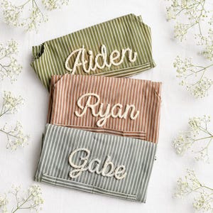 Könnte beinhalten: Drei personalisierte Stoffartikel mit Streifen in Olivgrün, Rost und Grau, jeweils mit einem Namen, der in einer seilartigen Schriftart gestickt ist: "Aiden", "Ryan" und "Gabe". Die Artikel sind auf einer weißen Oberfläche angeordnet, mit weißen Blumen verstreut.