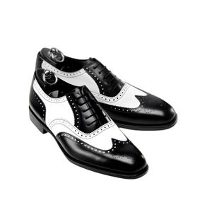 May include: A pair of two-tone wingtip dress shoes. The shoes are black with white accents on the sides and toe. The shoes have a classic lace-up design and are made of leather.