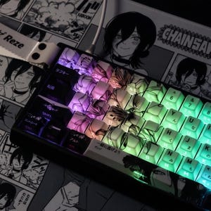 May include: A black mechanical keyboard with custom keycaps featuring anime artwork and Japanese characters. The keys are backlit with a gradient of colors, including purple, green, and white. A wrist rest and a lanyard with the name "Reze" are also visible.