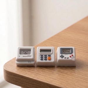 Può includere: Tre dispositivi elettronici bianchi in miniatura: un computer con una faccia sorridente, una calcolatrice con pulsanti arancioni e una console di gioco con uno schermo e pulsanti rossi. Sono disposti su una superficie di legno.