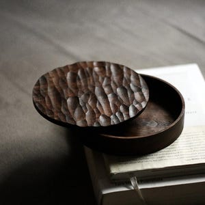 Puede incluir: Una caja de madera marrón oscuro con una tapa texturizada, parcialmente abierta. La tapa tiene un patrón tallado y la caja está encima de una pila de libros. La veta de la madera es visible y la estética general es rústica.