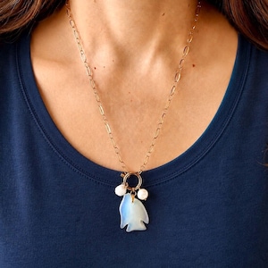 Può includere: Una collana a catena color oro con un ciondolo a forma di pesce. Il ciondolo è di colore azzurro chiaro e ha due accenti di perle bianche. La collana è indossata su una maglietta blu scuro.
