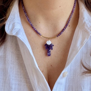 Può includere: Una collana di perline composta da piccole perline rotonde di ametista viola infilate su un cordino. Il centro include una barra metallica dorata curva, un piccolo ciondolo a forma di conchiglia bianca e un pendente in ametista intagliata.